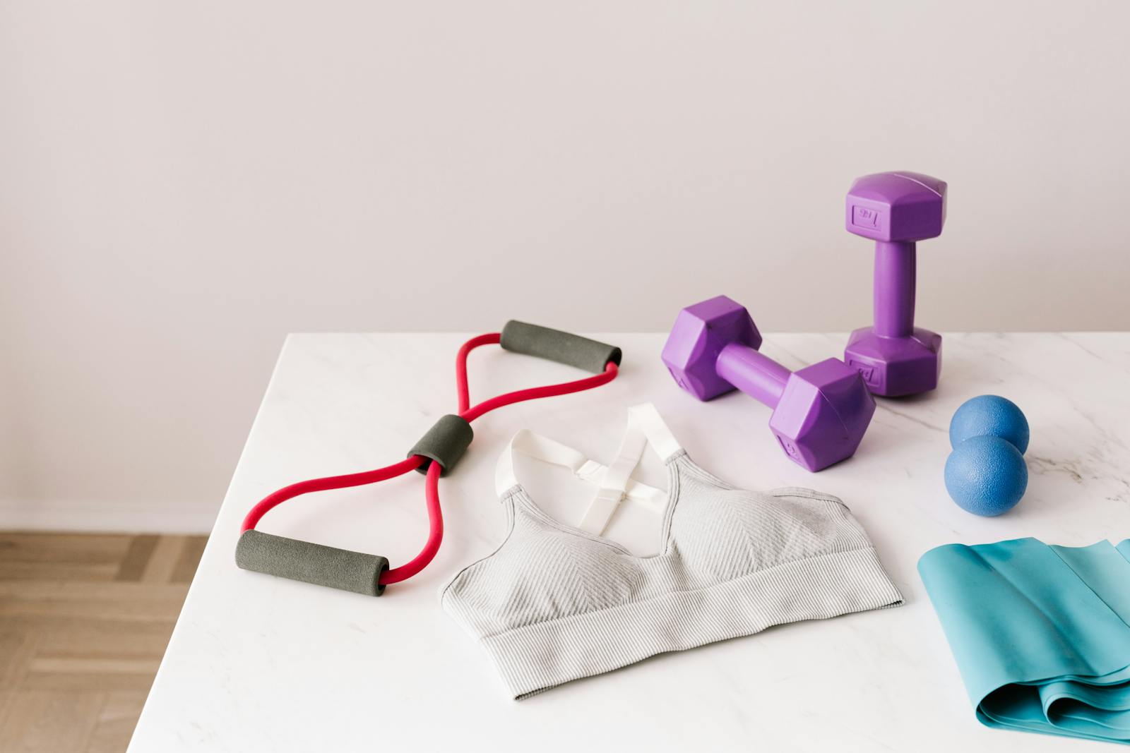 From above of white sports bra surrounded by purple dumbbells and blue tape expander and red tubular expander and blue massage double ball placed on marble surface of table